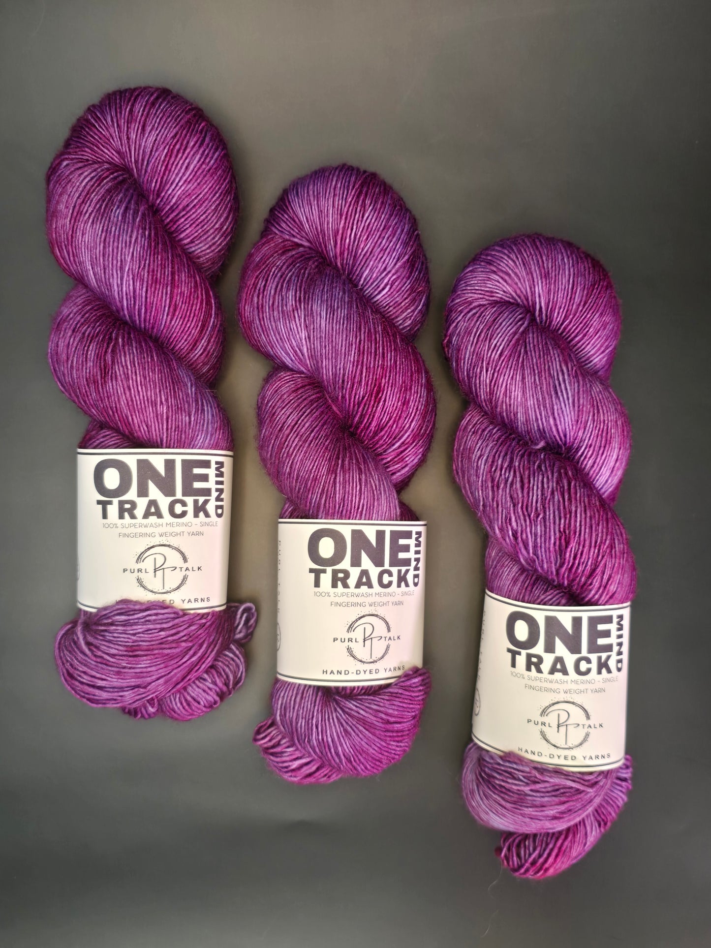 Three skeins of purple yarn with 'One Track Mind' labels on a gray background